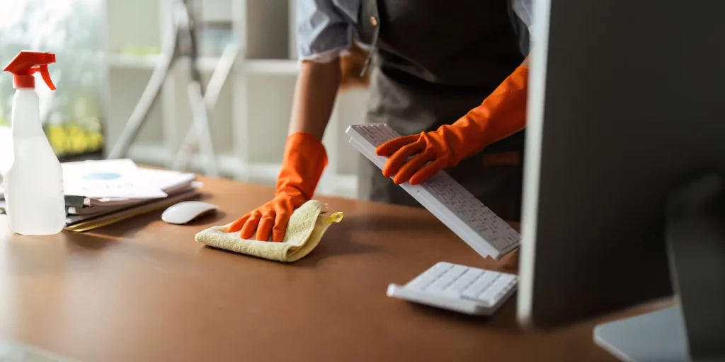 A person wearing orange gloves cleans a desk with a cloth, holding a keyboard. A computer monitor, mouse, and spray bottle are visible. The scene suggests office cleaning.