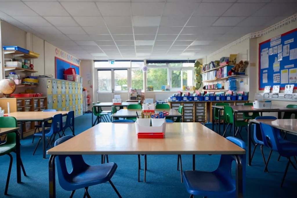 An empty classroom with several tables and colorful chairs. Shelves with books and educational materials line the walls. Large windows at the back let in natural light, and bulletin boards display various posters and student work.