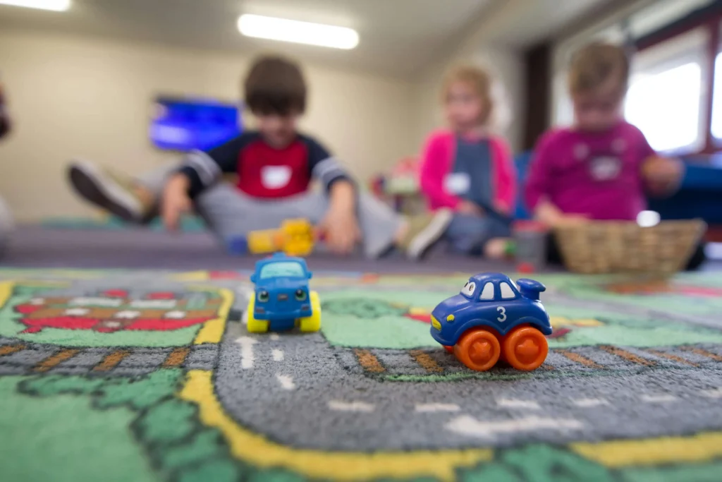 Two toy cars are on a play mat with roads in focus, while three young children sit in the blurred background, playing together indoors.